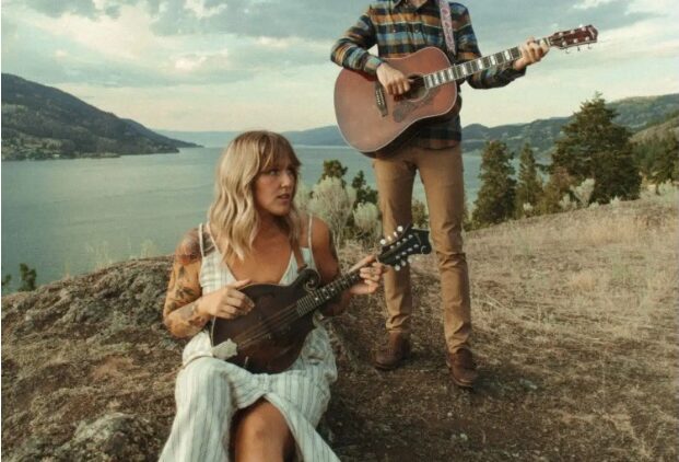 A woman in a striped dress sits on a rock playing a mandolin, while a man in a plaid shirt and cap stands behind her playing a guitar. They are outdoors, overlooking a scenic lake and hills under a cloudy sky.