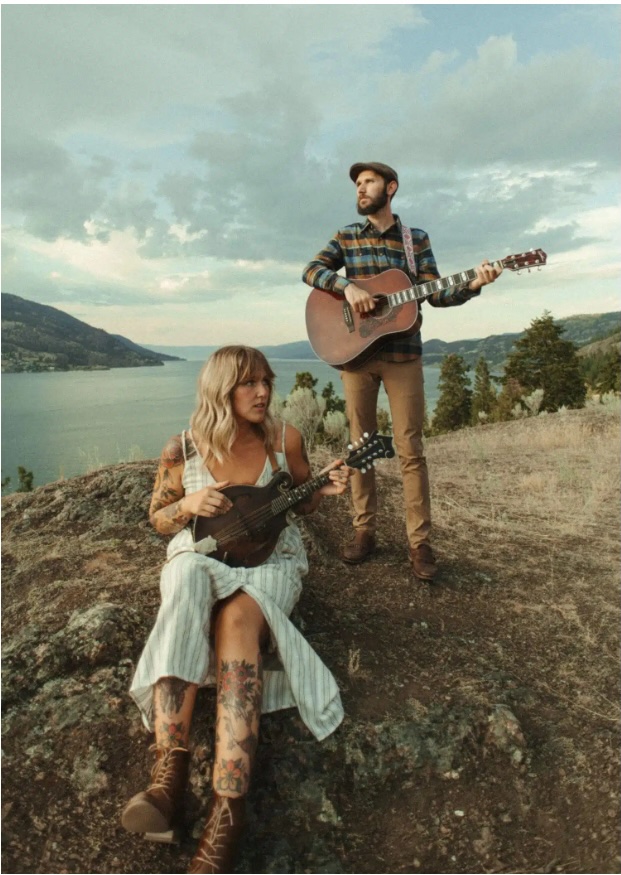 A woman in a striped dress sits on a rock playing a mandolin, while a man in a plaid shirt and cap stands behind her playing a guitar. They are outdoors, overlooking a scenic lake and hills under a cloudy sky.