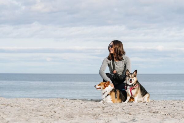 A woman wearing sunglasses kneels on a sandy beach with two corgi dogs, one on each side of her. The ocean and a cloudy sky are in the background.