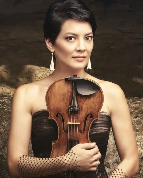 A woman with short dark hair and dangling earrings holds a violin upright in front of her chest. She wears a strapless dress and fishnet gloves, and looks confidently at the camera against a neutral background.