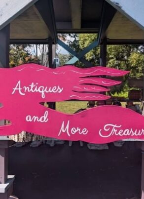 A pink mermaid-shaped sign reads Antiques and More Treasures and hangs from a wooden structure, with trees and outdoor items visible in the background.