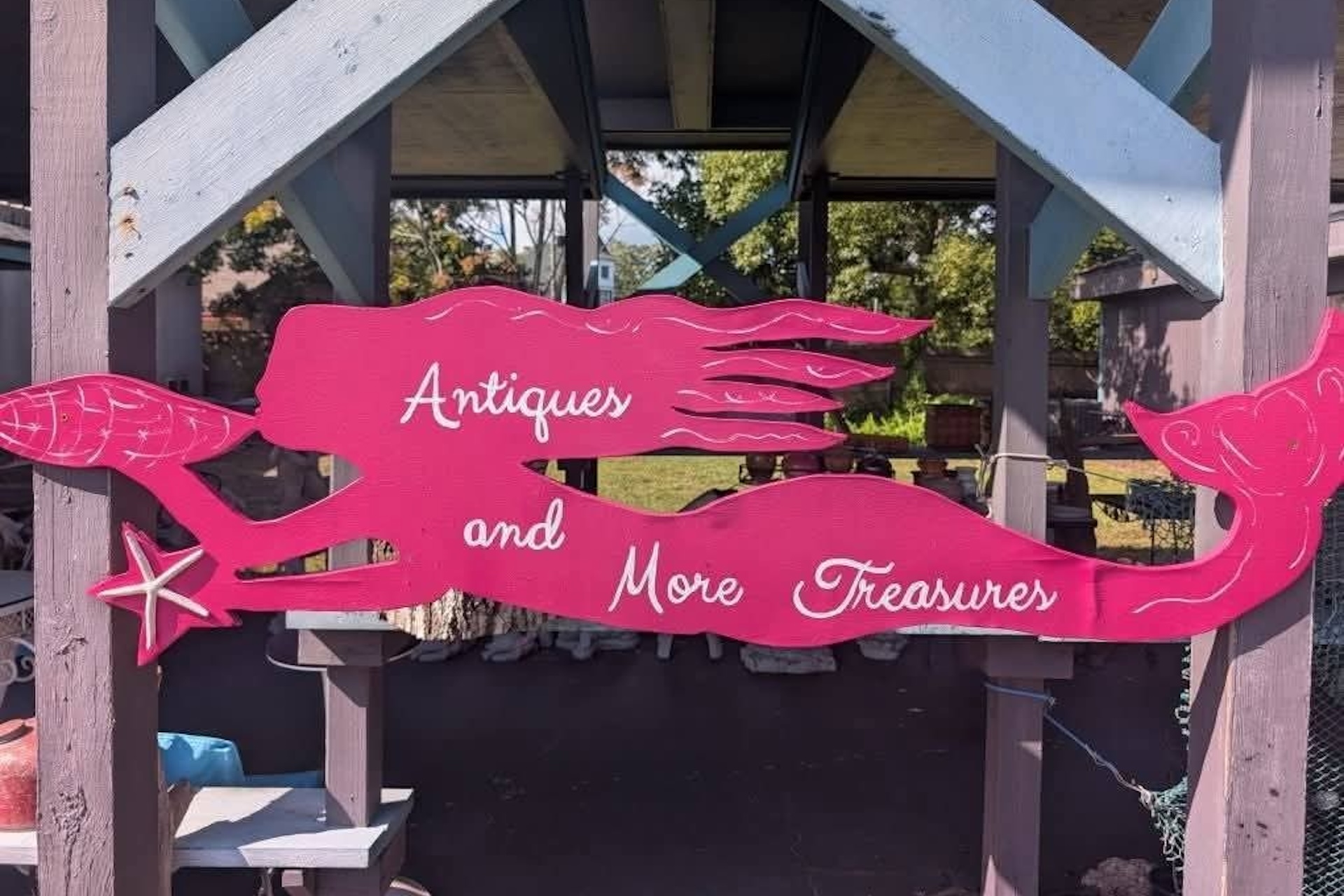 A pink mermaid-shaped sign reads Antiques and More Treasures and hangs from a wooden structure, with trees and outdoor items visible in the background.