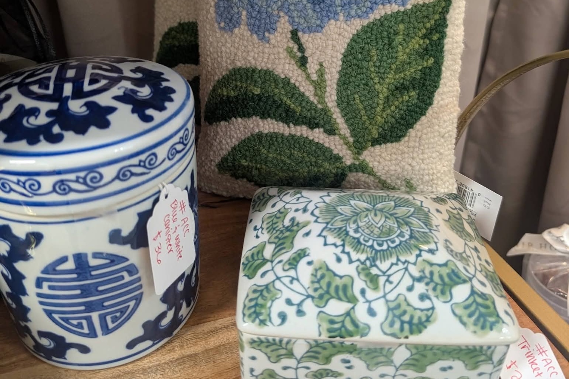 Blue and white ceramic jar, green and white patterned ceramic box, and a pillow with a blue and green floral design on a wooden surface. Red and white price tags are attached to the ceramics.