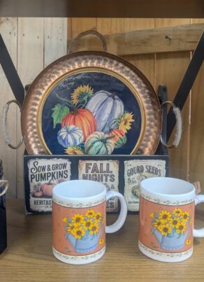 A shelf displays fall-themed decor: two blue glass jars with rope bows, a tray with pumpkins, two mugs with yellow flowers, an artwork of yellow flowers in a vase, and vintage seed packets, all against a wooden background.