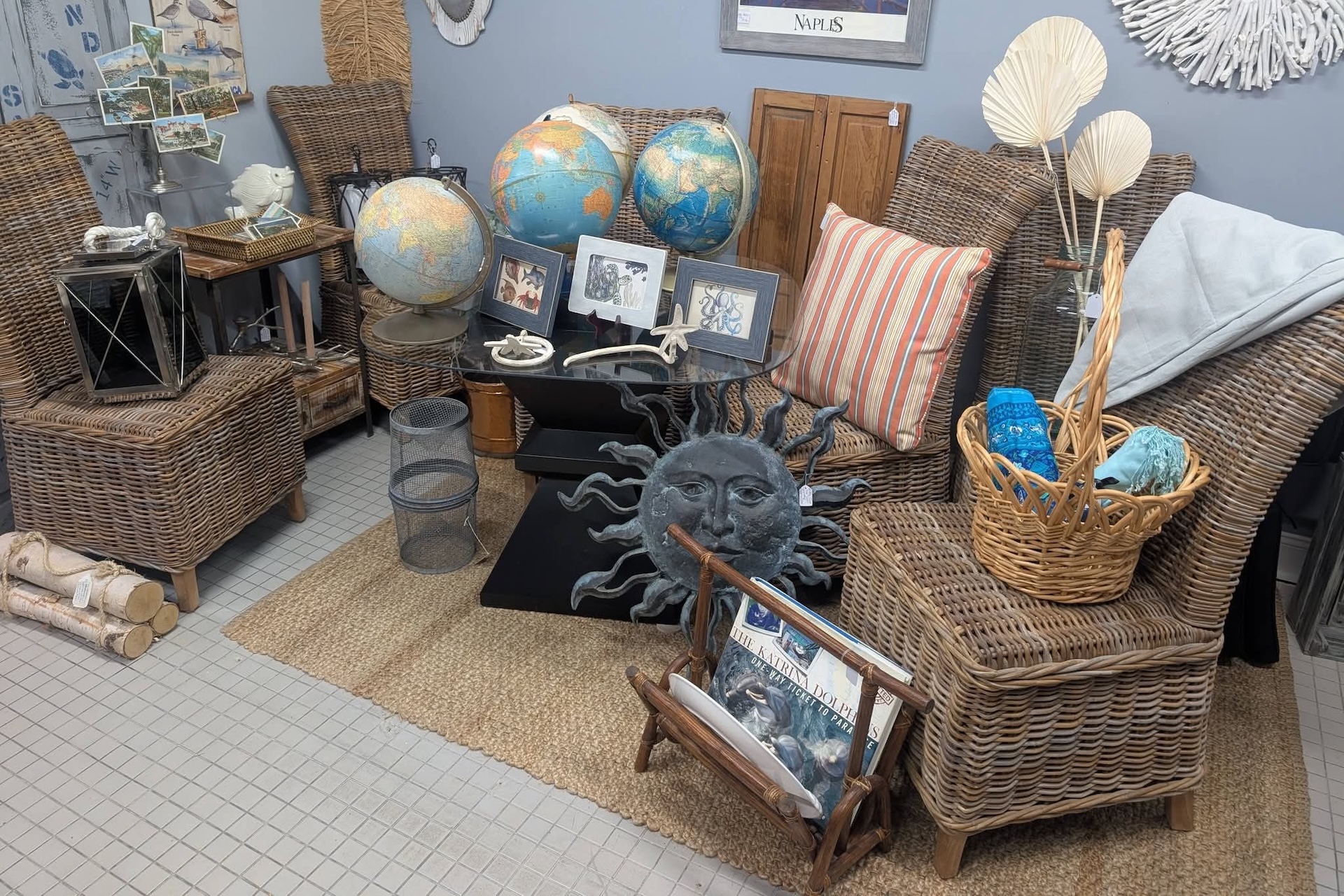 A cozy room with wicker chairs, globes, framed photos, a sun face decor, baskets, a lantern, and nautical-themed accents on a tiled floor.