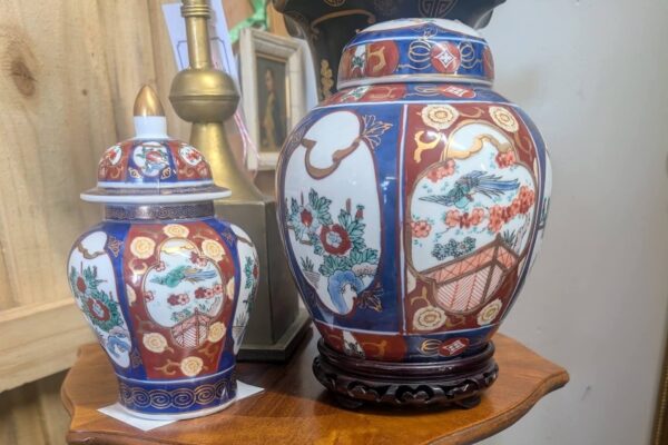 Two ornate, multicolored ceramic jars with intricate floral and landscape patterns sit on a wooden table. One jar has a lid; the other stands on a small carved wooden base. Both are decorated with blue, red, and gold accents.