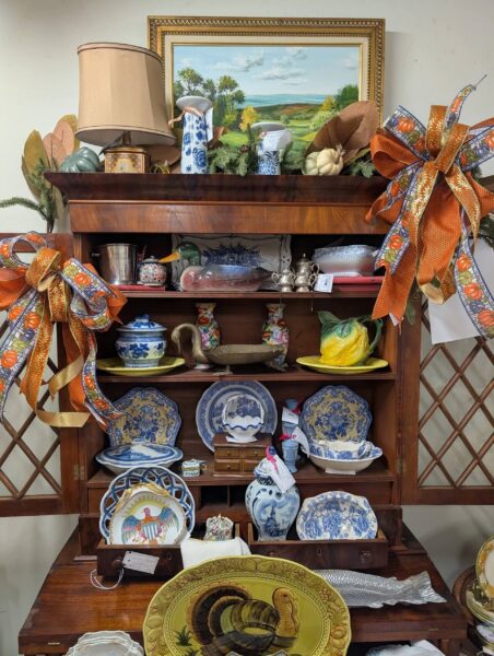 A wooden hutch displays blue and white china, a yellow ceramic fruit, turkey plates, vases, and decorative bows. A framed landscape painting and lamp sit on top. Autumn-themed decorations are visible throughout.