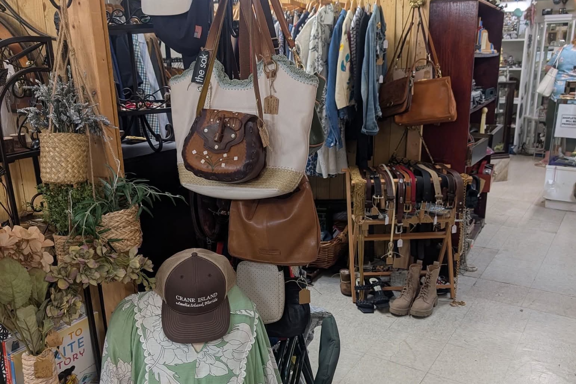A small shop displays purses, hats, belts, and boots. Handbags hang on racks, and various accessories are arranged on shelves. The scene is cozy and cluttered with vintage and rustic decor.