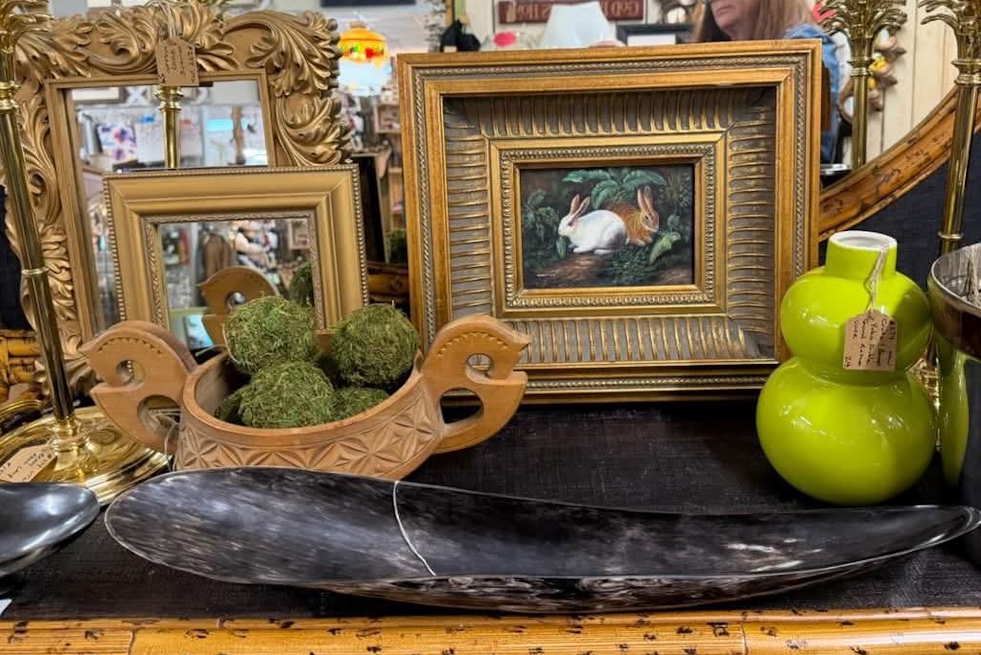 A tabletop with framed art, a wooden bowl holding green moss balls, a bright green vase, and a dark polished horn, all surrounded by ornate gold frames and mirrors in an antique store.