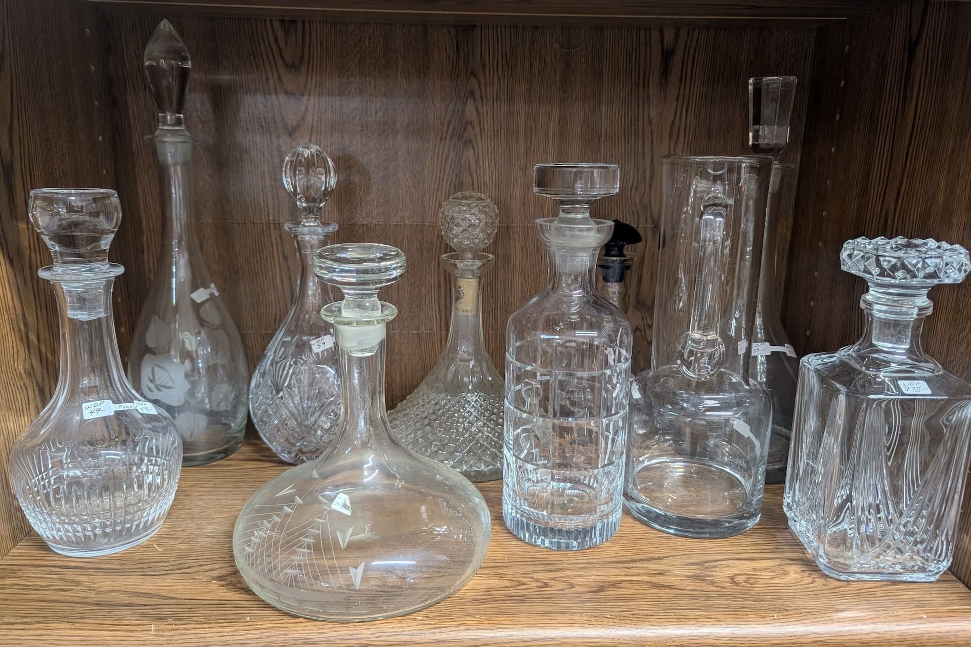 A collection of assorted clear glass decanters with various shapes and stoppers displayed on wooden shelves. Each decanter has a unique design and cut pattern.
