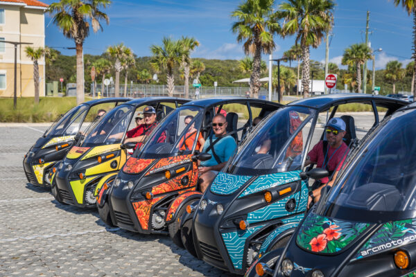 A row of five people sit smiling in colorful, three-wheeled electric vehicles parked outdoors on a sunny day, with palm trees and a building in the background.