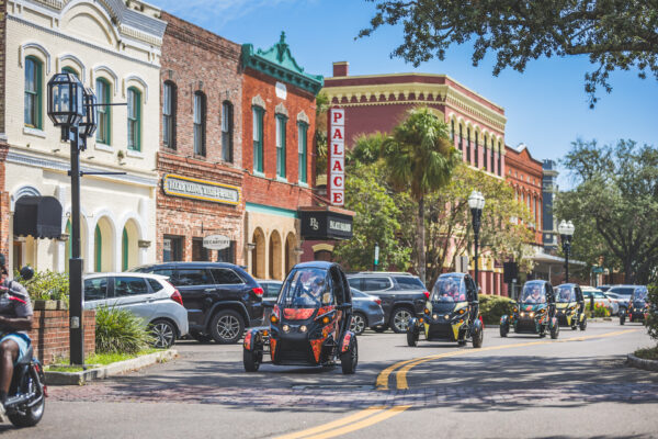 Several small three-wheeled electric vehicles drive down a sunny street lined with historic brick buildings, parked cars, and palm trees, with a clear blue sky overhead.