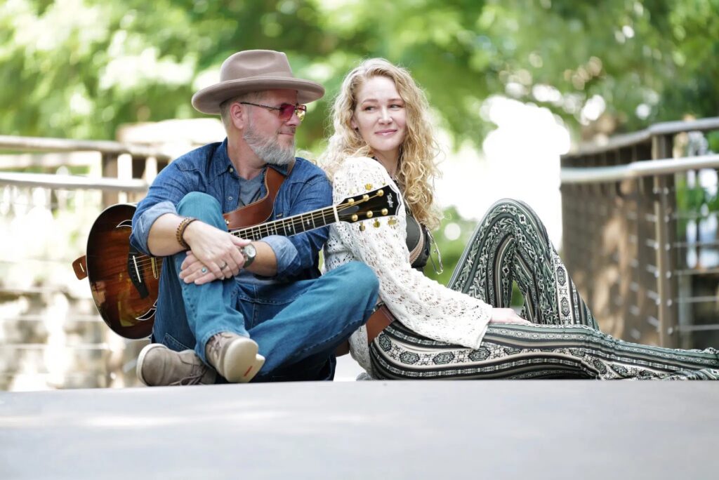 A man with a guitar and a woman sit back-to-back on a walkway, smiling. The background is green and bright, with trees and railing visible. Both appear relaxed and casual.