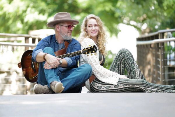 A man with a guitar and a woman sit back-to-back on a walkway, smiling. The background is green and bright, with trees and railing visible. Both appear relaxed and casual.