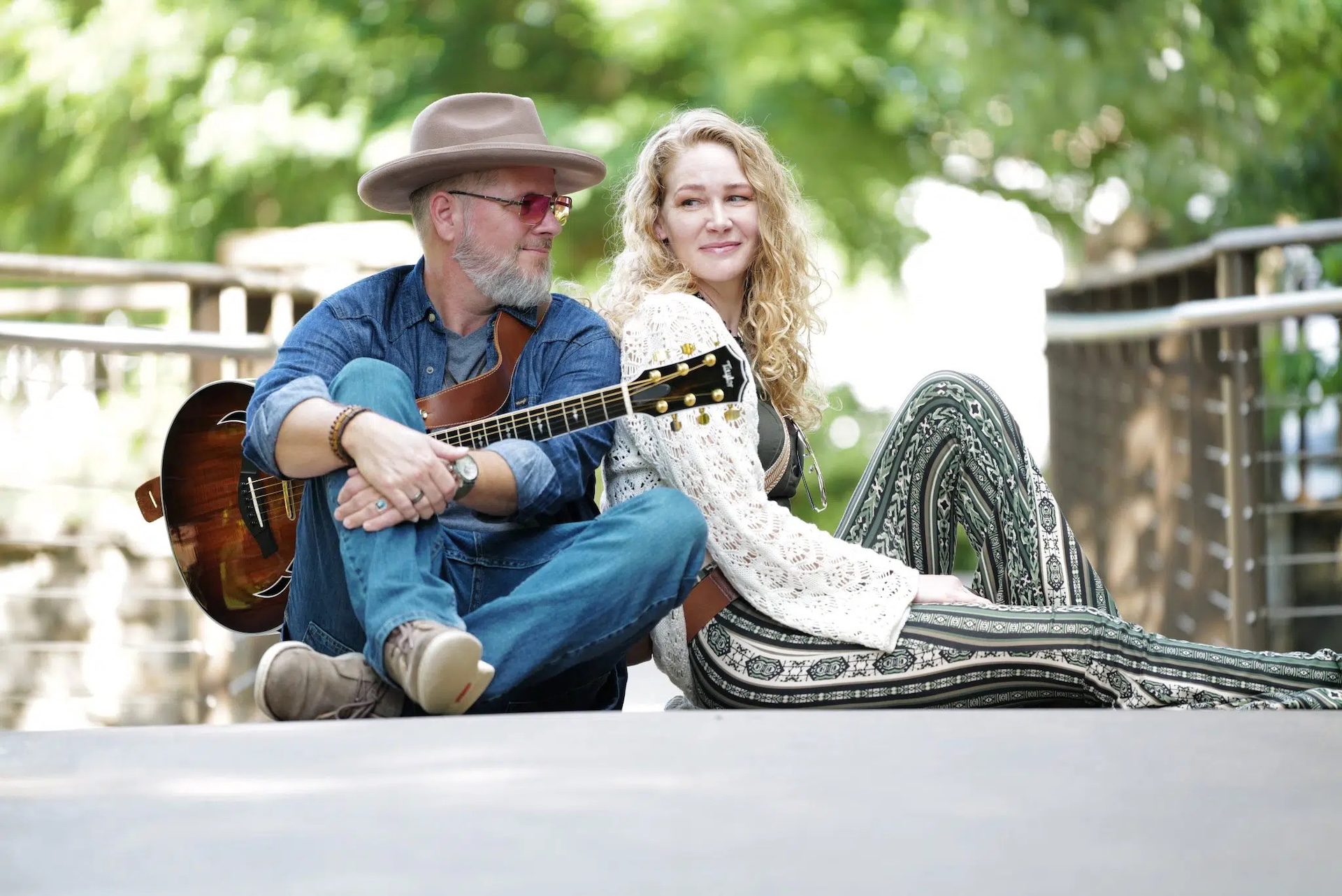 A man with a guitar and a woman sit back-to-back on a walkway, smiling. The background is green and bright, with trees and railing visible. Both appear relaxed and casual.