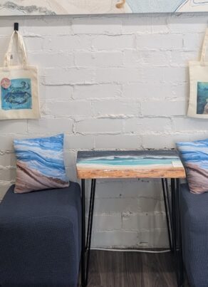 Two blue cushioned chairs face a small table with an ocean design. Art-themed tote bags hang on a white brick wall above, and two beach-themed pillows are on the chairs. A shelf is partially visible on the left.