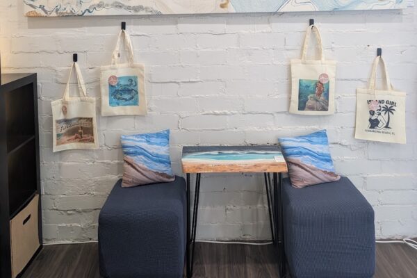 Two blue cushioned chairs face a small table with an ocean design. Art-themed tote bags hang on a white brick wall above, and two beach-themed pillows are on the chairs. A shelf is partially visible on the left.