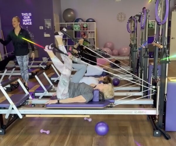 Three women are exercising on reformer Pilates machines in a bright studio with purple decor. An instructor stands at the front, guiding them. Exercise balls and equipment are visible, and a sign reads, This is our happy place.