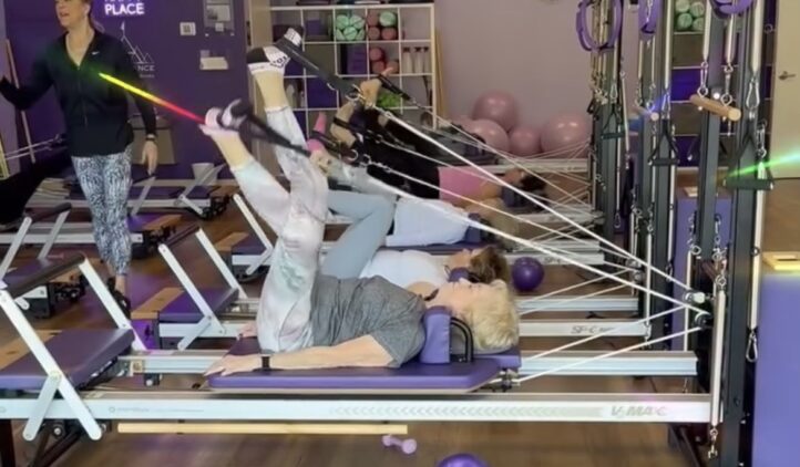 Three women are exercising on reformer Pilates machines in a bright studio with purple decor. An instructor stands at the front, guiding them. Exercise balls and equipment are visible, and a sign reads, This is our happy place.