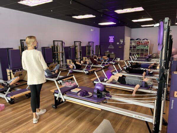 A fitness instructor stands at the front of a studio, observing a group of people lying on Pilates reformer machines, exercising in a spacious, purple-accented room with wooden floors and various fitness equipment.