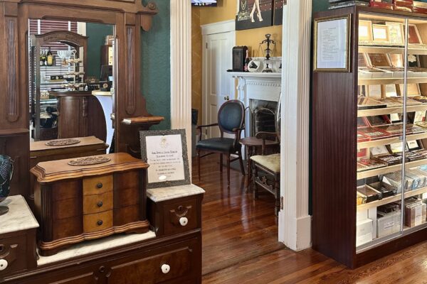 A cozy, vintage-style cigar lounge with wood floors, classic furniture, a humidor cabinet filled with cigars, and a fireplace in the background. A sign is displayed on a wooden counter with a mirror.