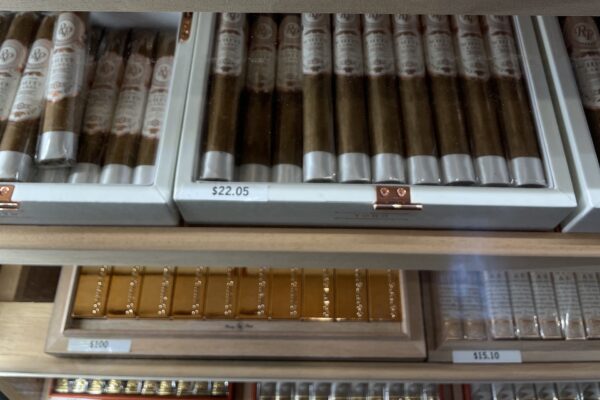 Boxes of cigars displayed on shelves in a store, each box labeled with prices. The top box is priced at $22.05, and others below are marked $15.10 and $1.00.