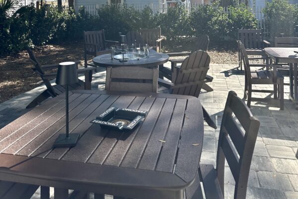 Outdoor patio with several wooden tables and chairs. A small black lamp and two copies of the board game Othello sit on the closest table. Sunlight creates shadows on the tiled ground; shrubs are visible in the background.