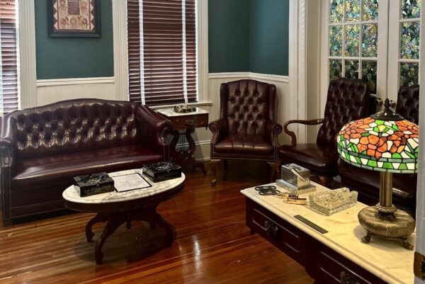 A vintage-style office with dark leather chairs, a matching sofa, wooden floors, a marble-topped coffee table, and a desk featuring a colorful stained glass lamp and office supplies.