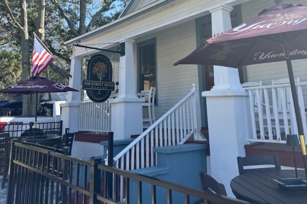 Outdoor seating area of a restaurant with tables, chairs, umbrellas, and an American flag. White wooden building with a porch, blue steps, and a sign reading Sun Street Grille near the entrance. Trees in the background.