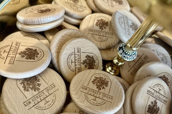 A pile of round wooden tokens engraved with Ash Street Cigar and a tree logo surrounds a gold pen with a jeweled holder.