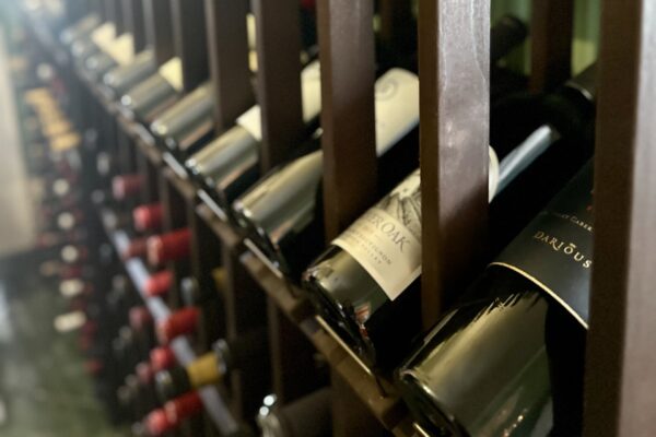 Close-up of wine bottles stored horizontally on a wooden rack, with a mix of red and white wine labels visible. The bottles are arranged in rows, and the focus is on the front labels.
