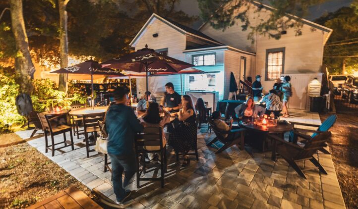 People gather at outdoor tables under large umbrellas on a patio at night, enjoying food and conversation. Warm lighting and trees surround the area, with a house in the background.
