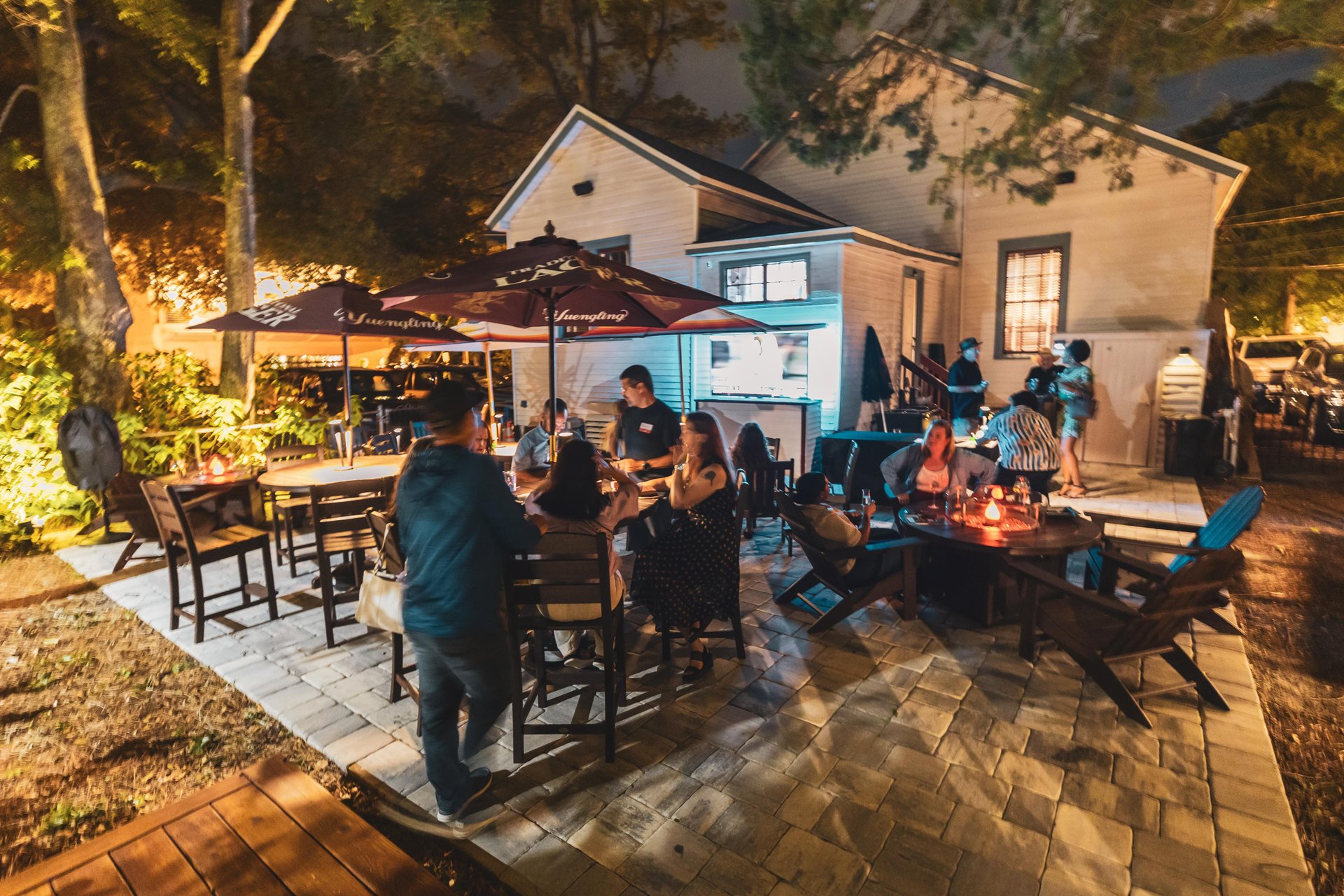 People gather at outdoor tables under large umbrellas on a patio at night, enjoying food and conversation. Warm lighting and trees surround the area, with a house in the background.