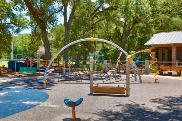 A playground with climbing nets, slides, and various play structures surrounded by trees and shaded areas. There is a building with a metal roof to the right and benches around the play area.