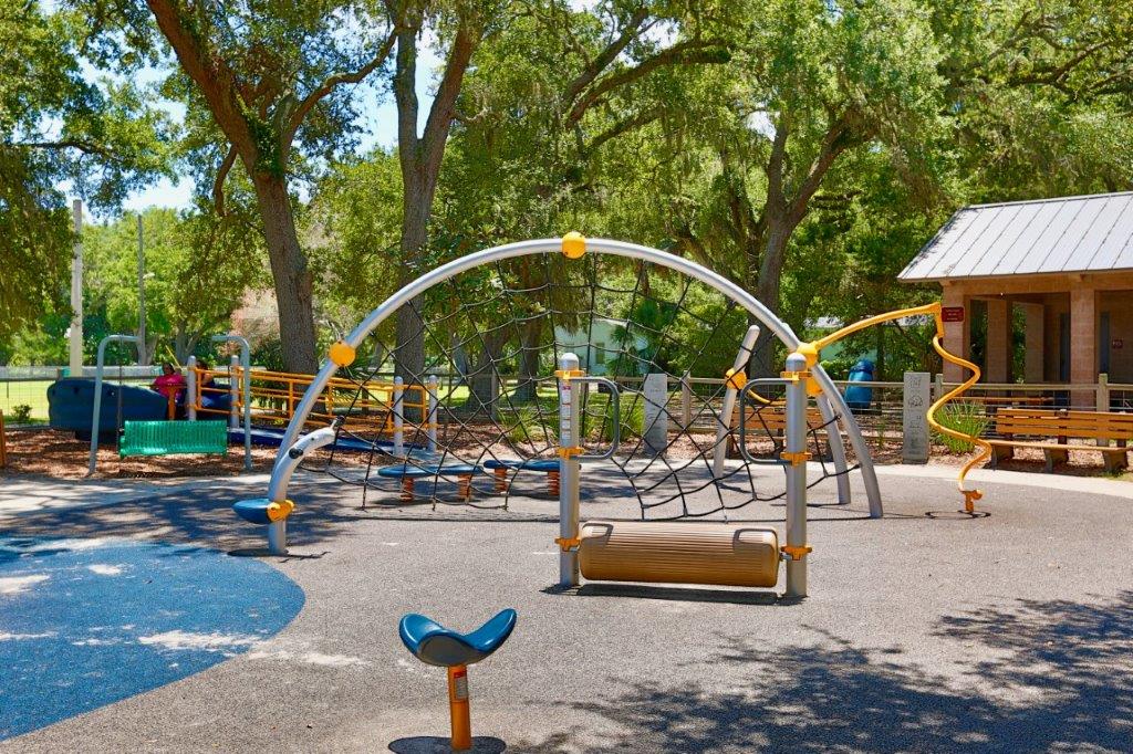 A playground with climbing nets, slides, and various play structures surrounded by trees and shaded areas. There is a building with a metal roof to the right and benches around the play area.