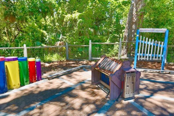An outdoor musical playground features colorful drum tubes, a large xylophone, and an oversized chime set, all surrounded by trees and a wooden fence under bright sunlight.