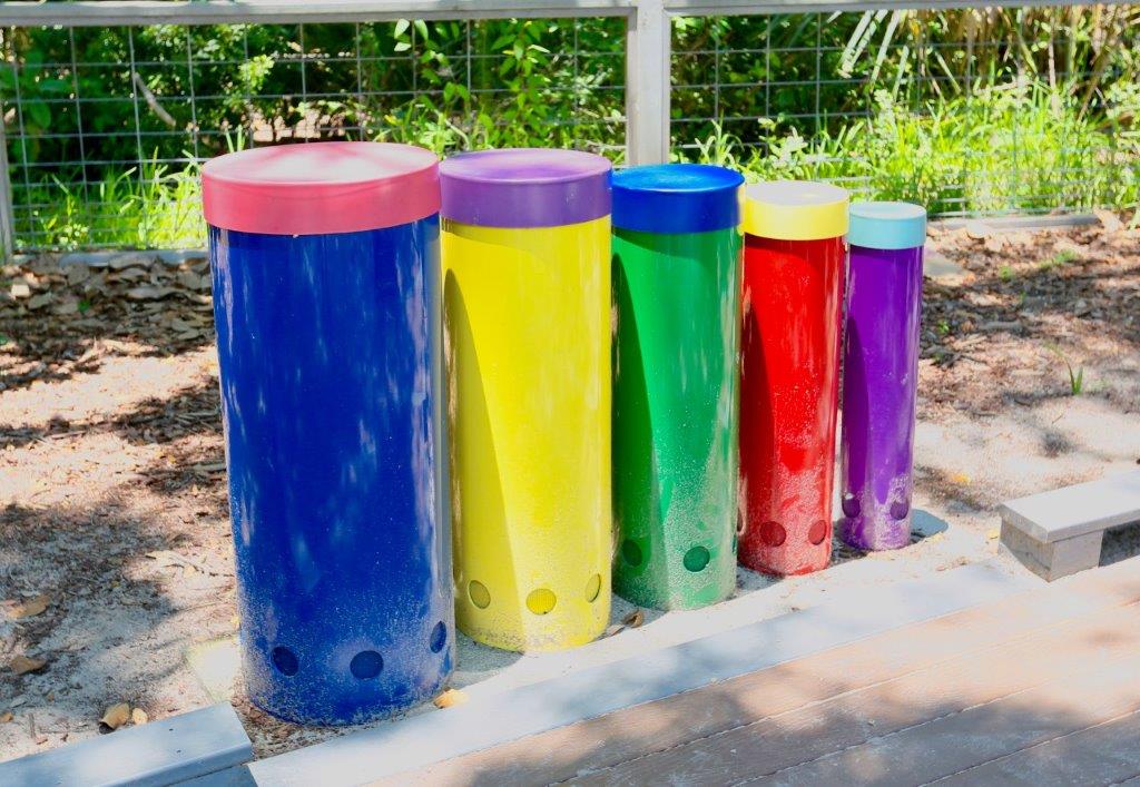Five large, colorful outdoor drums in blue, yellow, green, red, and purple stand in a row on a playground, surrounded by greenery and sunlight.