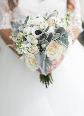 A bride in a white lace dress holds a bouquet of white roses, anemones, and dusty miller leaves.