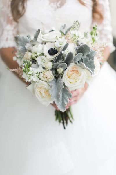 A bride in a white lace dress holds a bouquet of white roses, anemones, and dusty miller leaves.