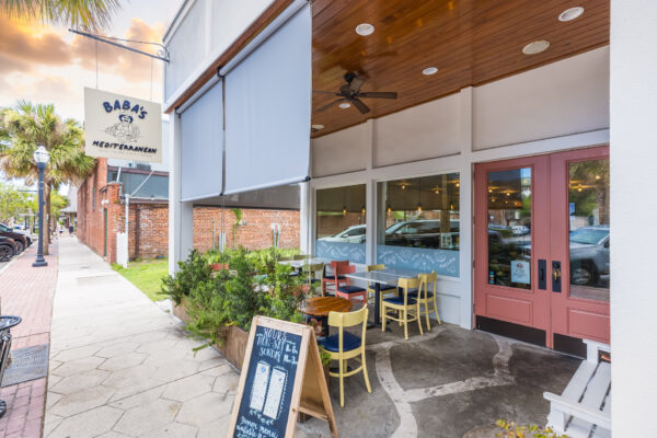 Outdoor seating area of a Mediterranean restaurant called Baba’s, with colorful chairs and tables, a chalkboard menu, and a wooden ceiling. The entrance has double pink doors and large windows, with plants nearby.