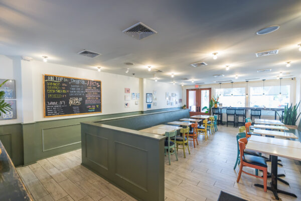 A bright, modern café with light wood floors, colorful chairs, and tables neatly arranged. A chalkboard menu hangs on the wall, and large windows let in natural light, creating an inviting atmosphere.