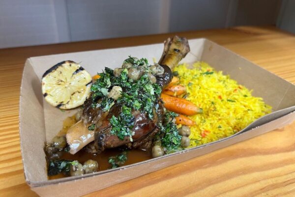 Braised lamb shank topped with herbs and capers, served with roasted baby carrots, yellow rice, and a grilled lemon half in a brown takeout container on a wooden surface.