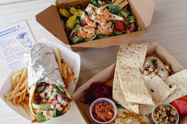 Three takeout boxes with Mediterranean food: a shrimp salad, a gyro wrap with fries, and a platter with pita, hummus, tabbouleh, veggies, and sauces, arranged on a light table.