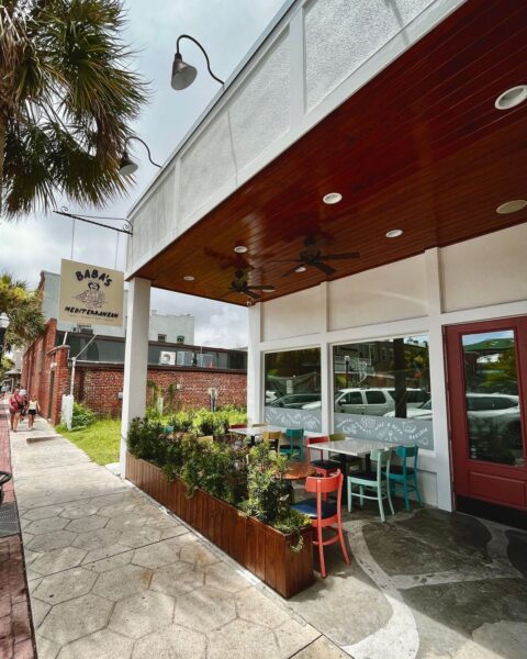 A sidewalk view of Baba’s Mediterranean restaurant with outdoor seating, colorful chairs, potted plants, and a red door. A palm tree and the restaurant’s sign are visible, with cloudy skies overhead.