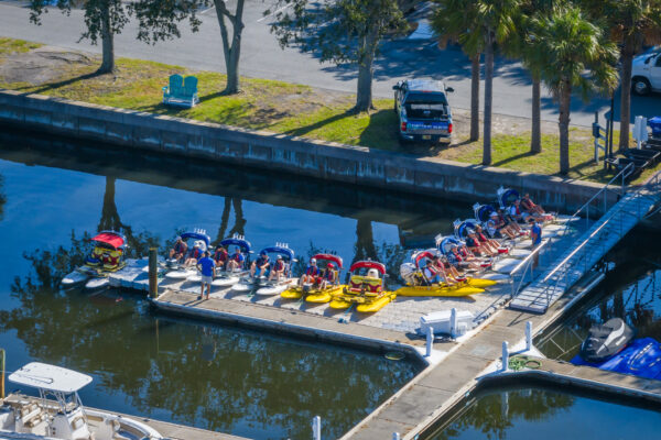 A dock lined with several yellow water bikes, each with a red or blue seat, sits on calm water next to a parking lot with trees, a parked truck, and empty green benches.