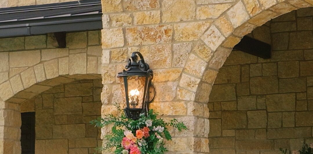 A rustic stone building features an arched entryway. A black lantern with a floral arrangement of pink, orange, and white flowers hangs on a stone pillar, with more flowers and wooden chairs visible under the arch.