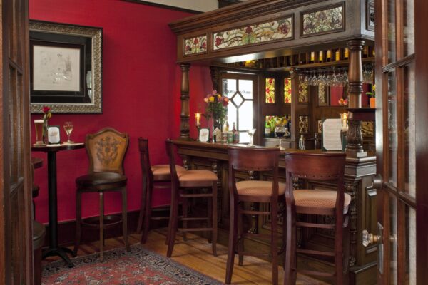 A cozy, vintage-style home bar with carved wood, stained glass panels, high-backed chairs, a red accent wall, framed art, flowers, and a patterned rug on a wooden floor.