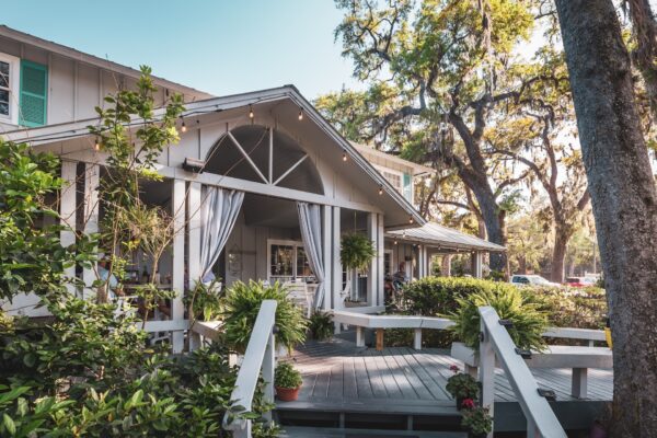 A charming house with a covered porch, white railings, hanging plants, and curtains, surrounded by lush greenery and large trees on a sunny day.