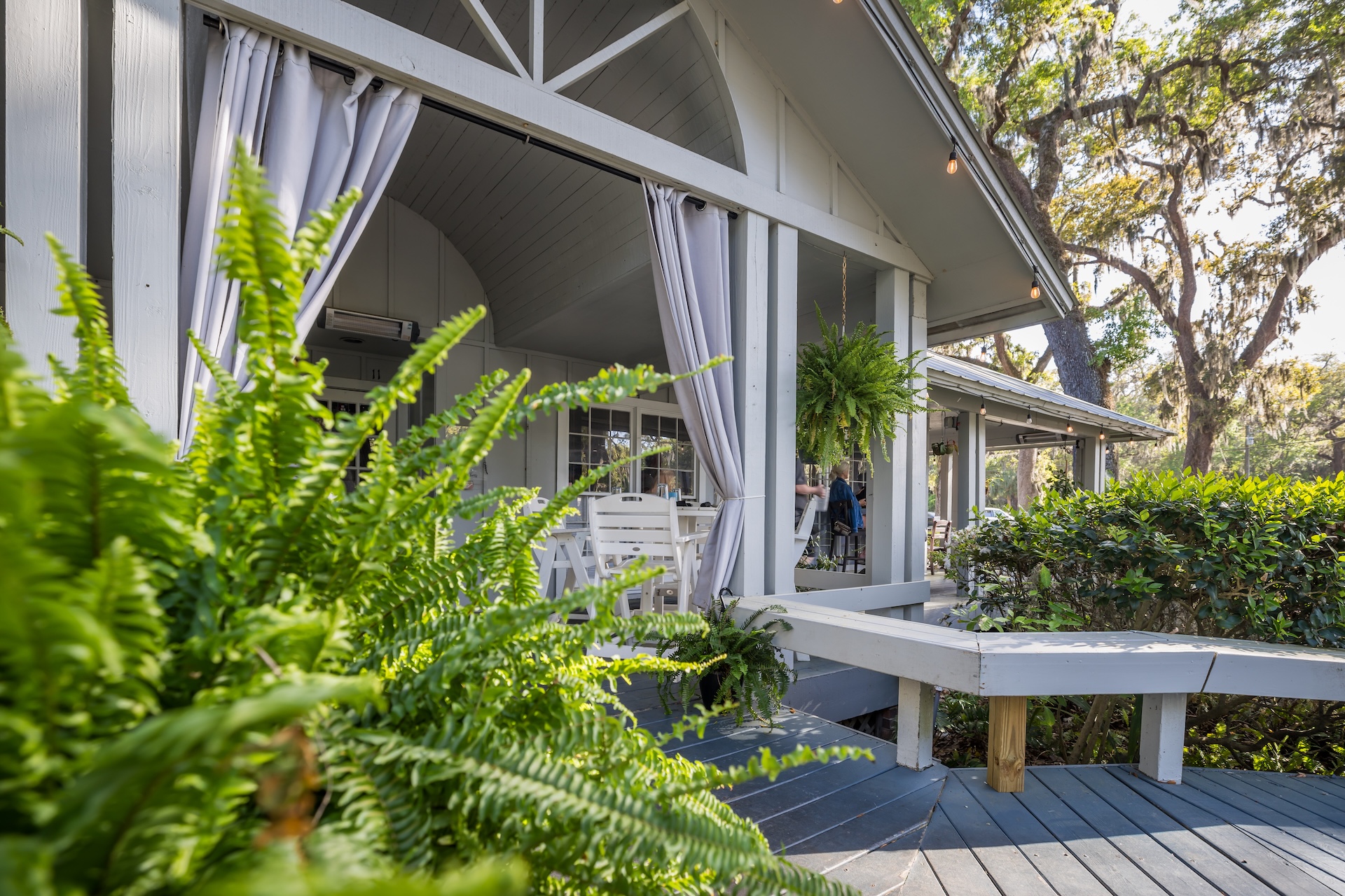 A cozy outdoor porch with white wooden benches and hanging plants, surrounded by greenery and trees. Light curtains hang from the porch, creating a relaxed and inviting atmosphere.