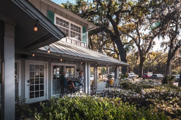 Two people sit at a table on the porch of a cozy, gray cottage-style café surrounded by lush greenery and large trees. Sunlight filters through the branches, and parked cars are visible in the background.